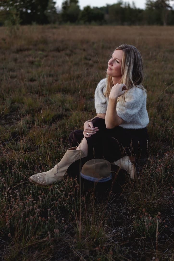 Jolanda zit in het gras op de heide en kijkt dromerig opzij, in zacht avondlicht